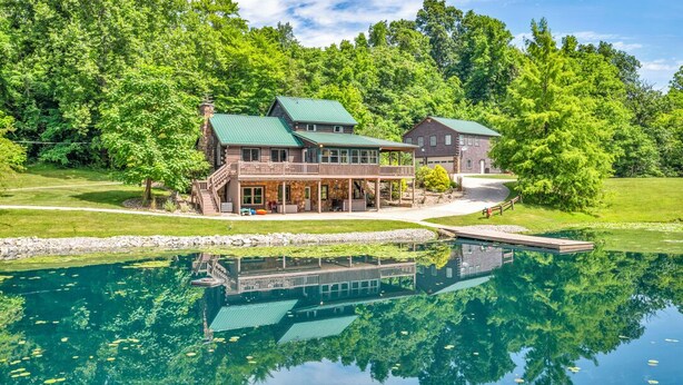 Une grande maison à deux étages, dotée d’un toit végétalisé, avec une jetée s’avançant dans un lac aux eaux limpides, le tout entouré d’une végétation luxuriante.