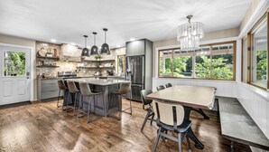 Une cuisine moderne dotée d’un îlot central, de tabourets de bar, d’une table à manger et de grandes fenêtres offrant une vue sur la verdure.