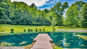 Une terrasse en bois mène à un étang aux eaux limpides, entouré d’une végétation luxuriante et d’arbres.