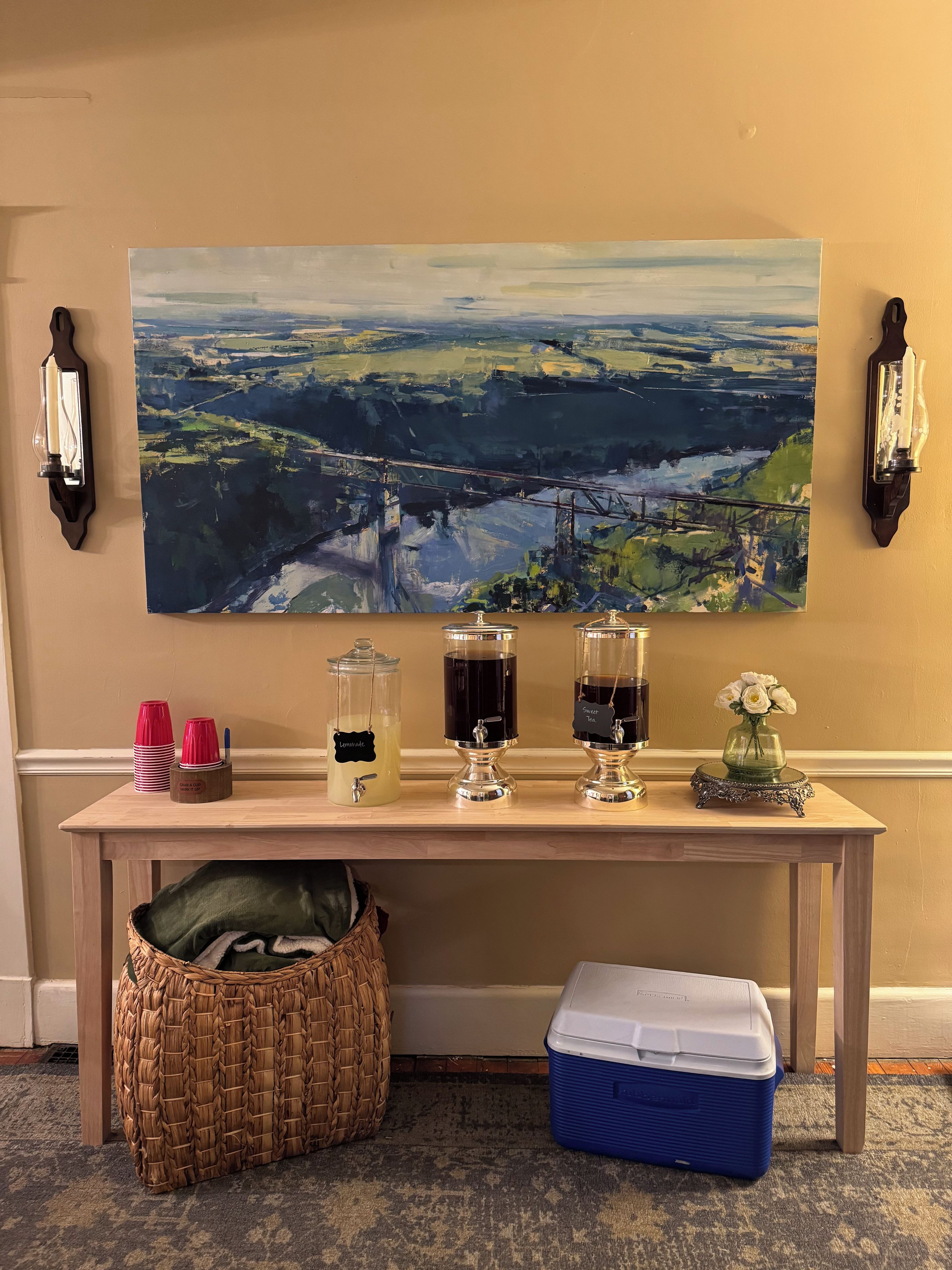 A room with a large landscape painting, a table with drink dispensers, a wicker basket, and a cooler.