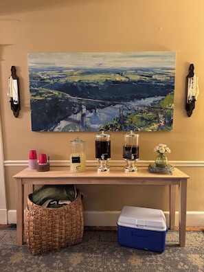 A room with a large landscape painting, a table with drink dispensers, a wicker basket, and a cooler.