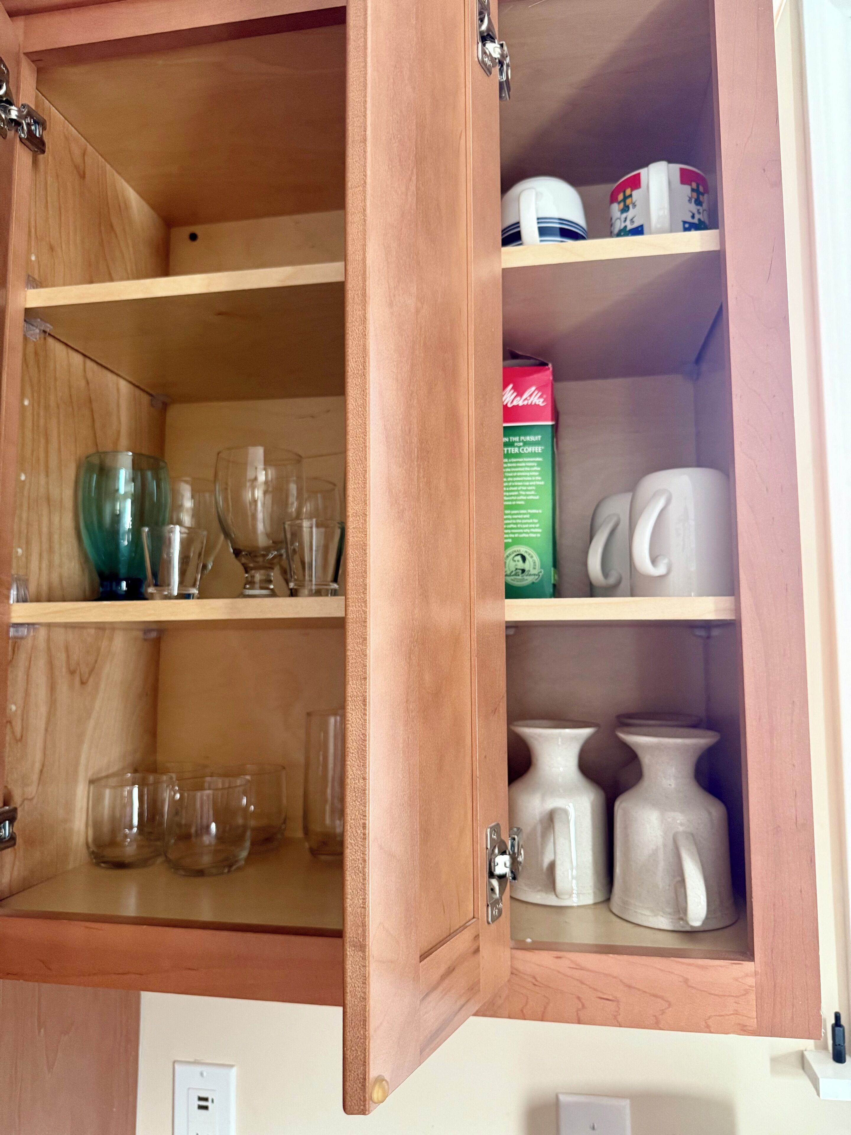 A wooden cabinet with shelves containing glassware, mugs, and a box of coffee.