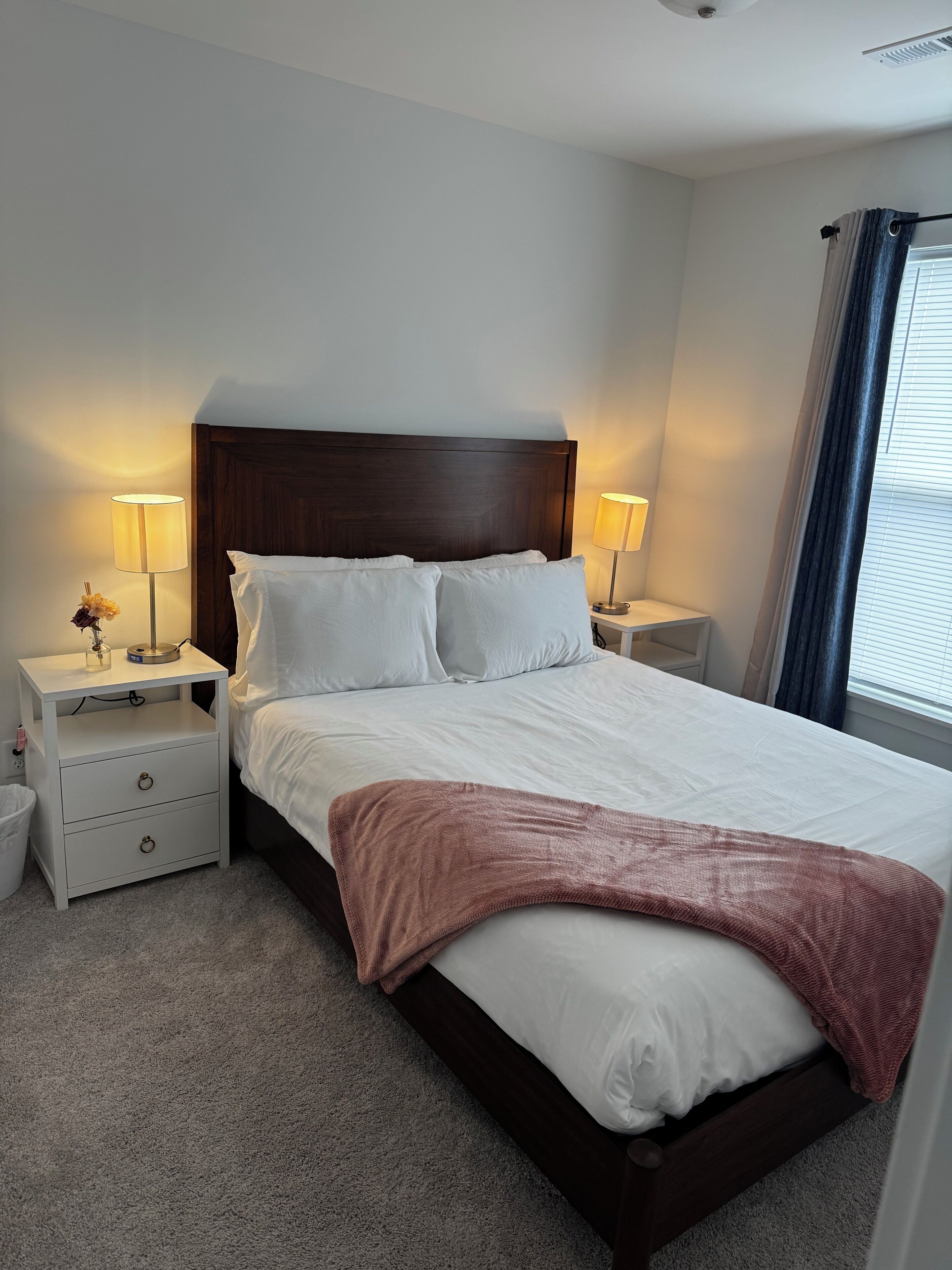 A neatly made bed with white linens and a brown throw blanket, flanked by two bedside tables with lamps, in a room with a wooden headboard and a window with curtains.