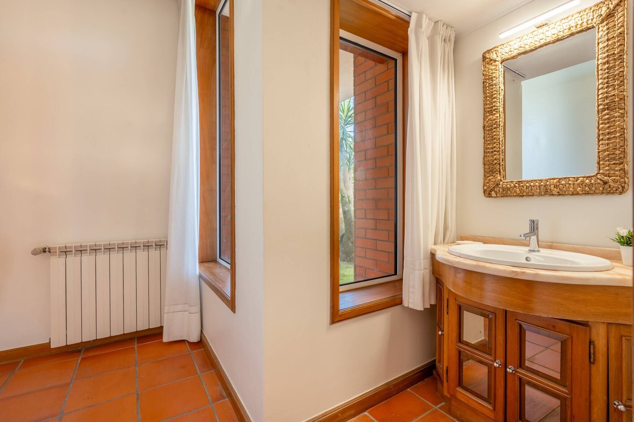 Een badkamer met een witte radiator, een raam met uitzicht op een bakstenen muur, een houten kastje met een wastafel en een spiegel met een geweven frame.
