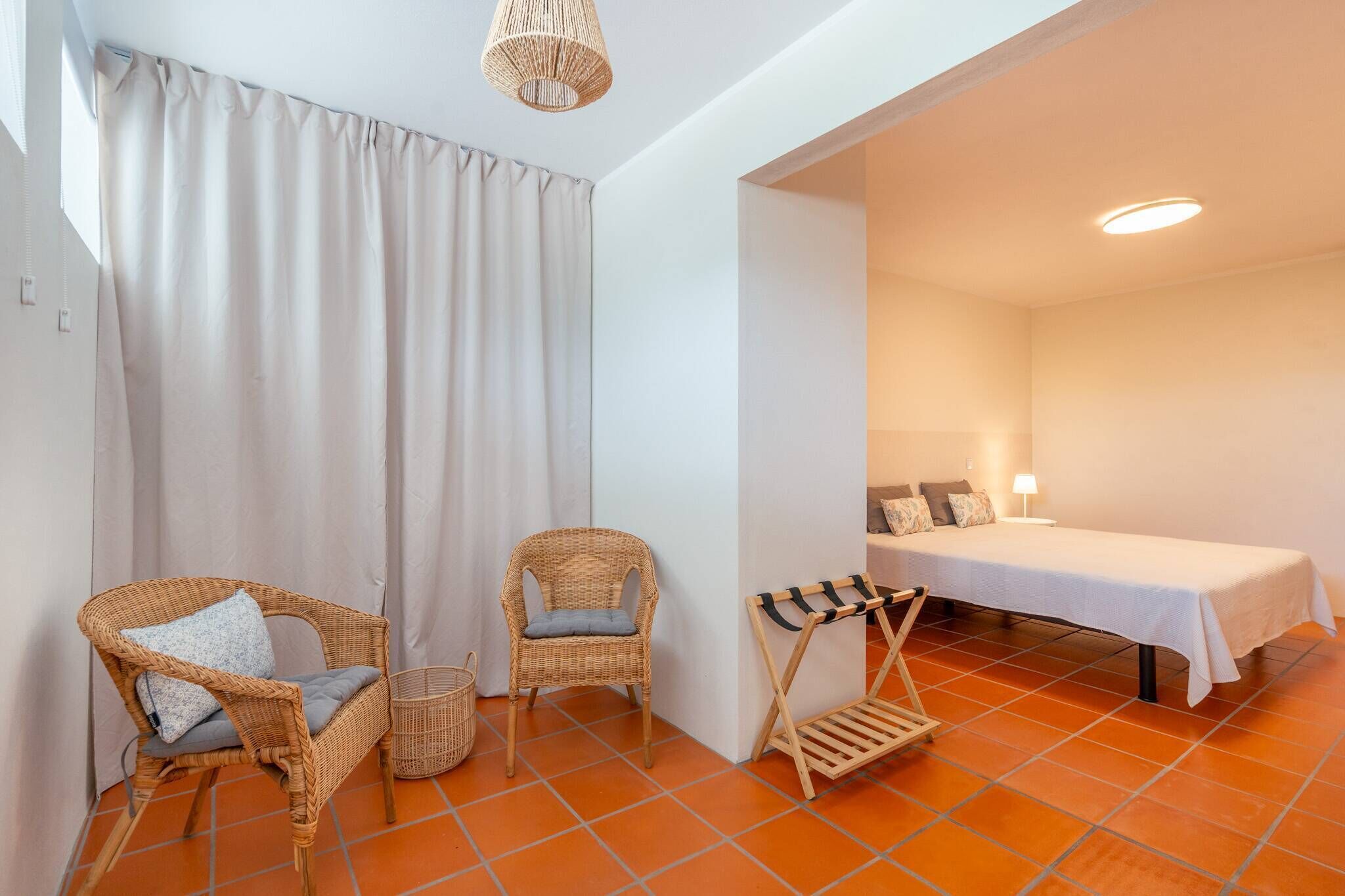 A room with a bed, two wicker chairs, a small table, and a hanging rack.