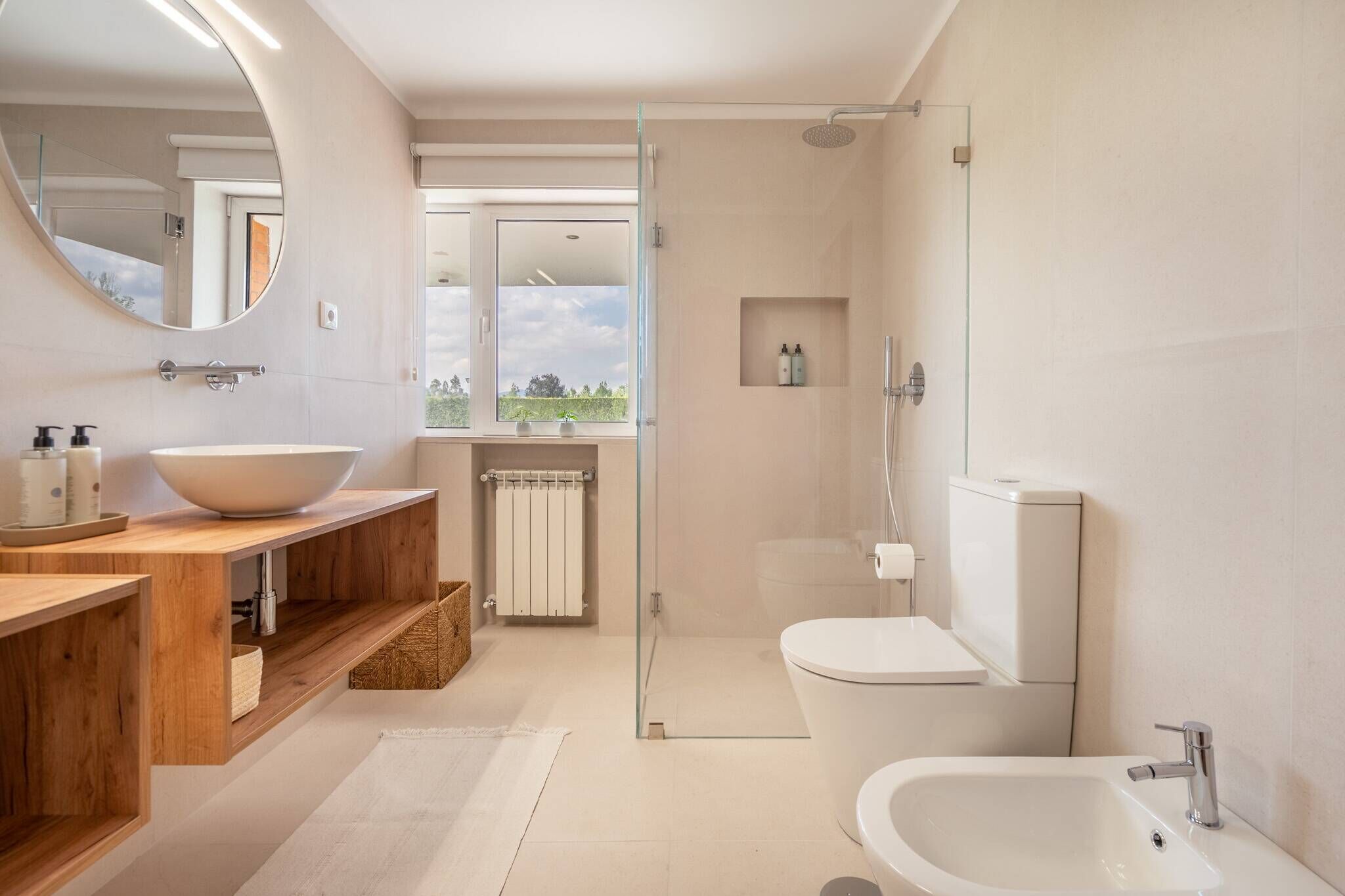 Un baño moderno con un espejo ovalado grande, un tocador de madera con lavabo, una bañera independiente y una zona de ducha con mampara de vidrio.