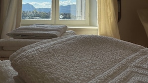 Ein Bett mit weißer, strukturierter Decke, ein Fenster mit Vorhängen und Blick auf Berge und Wolken.