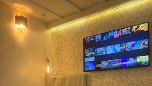 Ein modernes Zimmer mit einem runden Tisch, zwei Stühlen, einem an der Wand befestigten Fernseher, der verschiedene Sendungen zeigt, und einer dekorativen Leuchte.