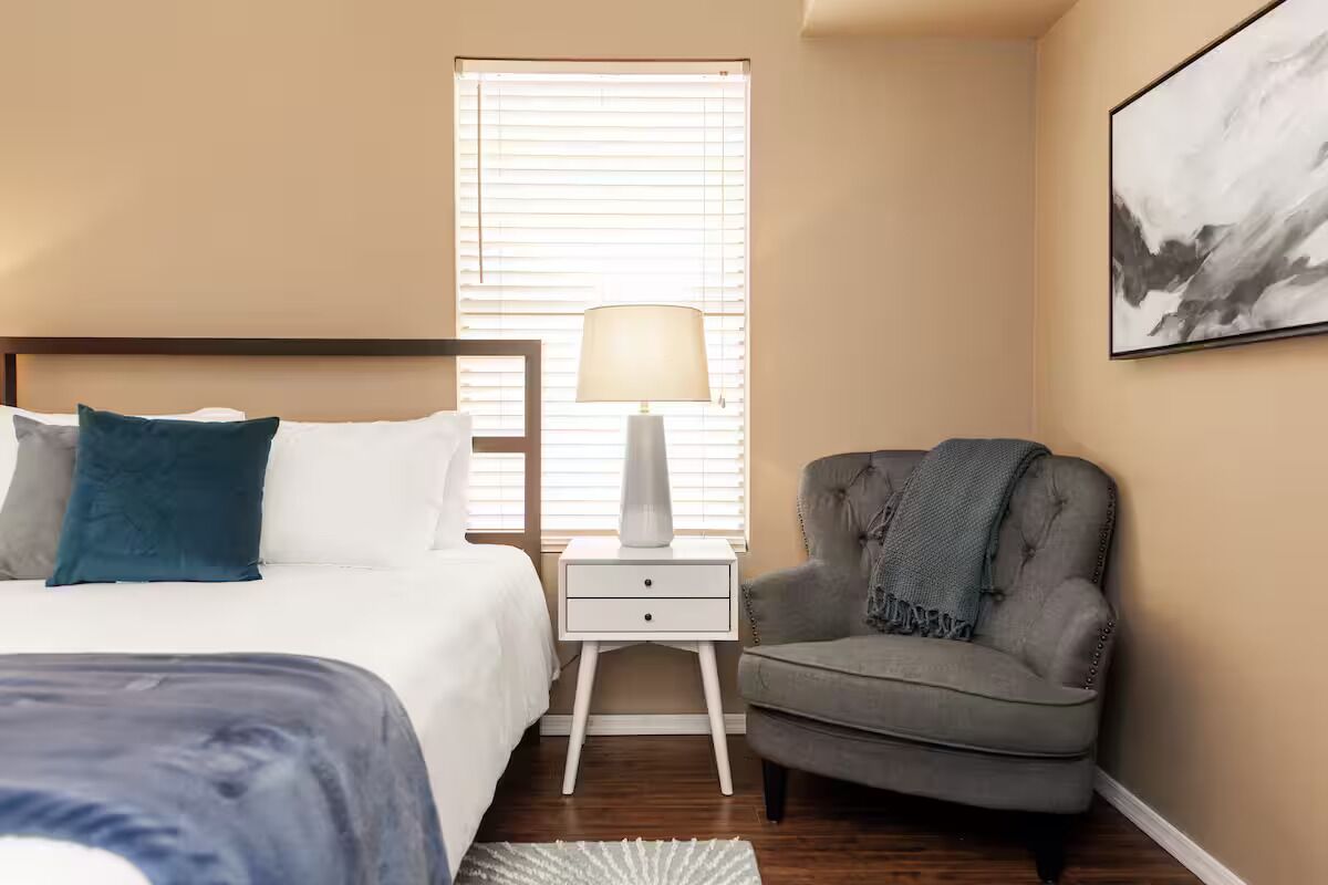A bedroom with a bed, bedside table, lamp, chair, and a painting on the wall.