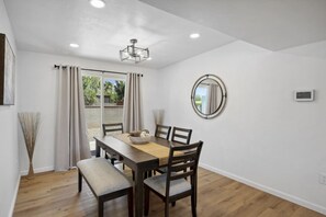 A dining area with a wooden table and chairs, a ceiling light, a round mirror, and a window with curtains. - Boho on Bermuda | Private Pool & Pet Friendly (Tucson)
