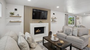 A modern living room with a flat-screen TV mounted above a fireplace, a grey sofa, and a coffee table with remote controls. - Boho on Bermuda with Private Pool in Tucson (Tucson)