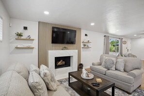 Ein modernes Wohnzimmer mit Flachbildfernseher über einem Kamin, einem grauen Sofa und einem Couchtisch mit Fernbedienungen.