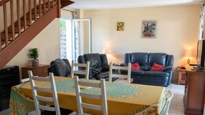 Un comedor con una mesa dispuesta para cuatro personas, rodeada de sillas blancas. Un sofá de cuero negro y un mueble de madera con una televisión se observan al fondo.