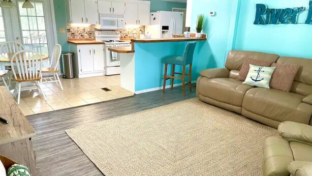 A living room with a beige sofa, a coffee table, and a rug. There is a kitchen area with white cabinets and a blue accent wall. - 3 BR 2 BA House w/Dock on Deep Canal in Bass Capital (Crescent City)