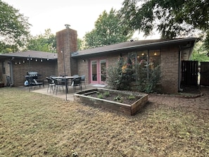 Een achtertuin met een huis van baksteen, een barbecue, een eettafel met stoelen en een verhoogd plantenbak.