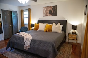 Een slaapkamer met een bed, nachtkastje, stoel, raam met gordijnen en een deur.