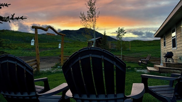 Un espacio al aire libre rústico con sillas Adirondack y una mesa de madera, con vistas a un paisaje cubierto de hierba donde se distingue una cabaña de madera y una colina al fondo.