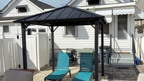 Un patio con zona de estar techada, dos sillones de relax, una mesa redonda y una chimenea, todo ello delimitado por una cerca blanca.