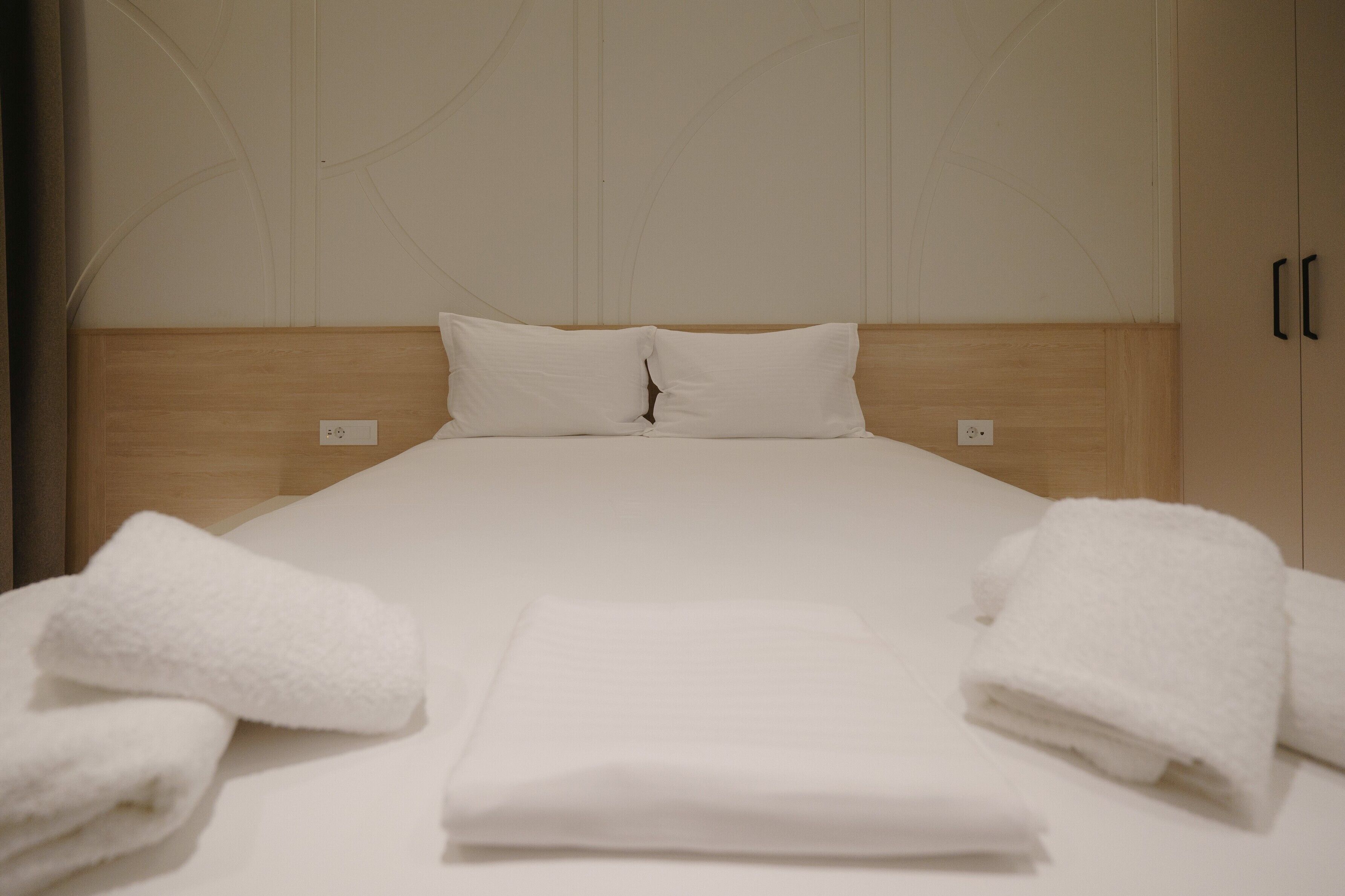 A neatly made bed with white linens and two pillows, a wooden headboard, and a plain wall with a subtle design.