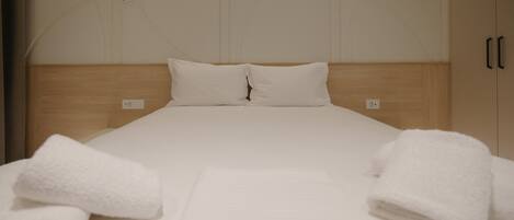A neatly made bed with white linens and two pillows, a wooden headboard, and a plain wall with a subtle design.
