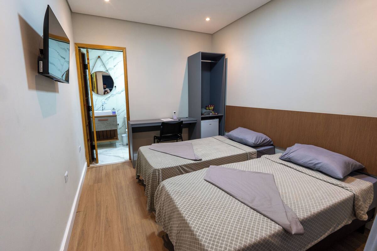 Quarto de hotel com duas camas, uma escrivaninha, um espelho e uma televisão.