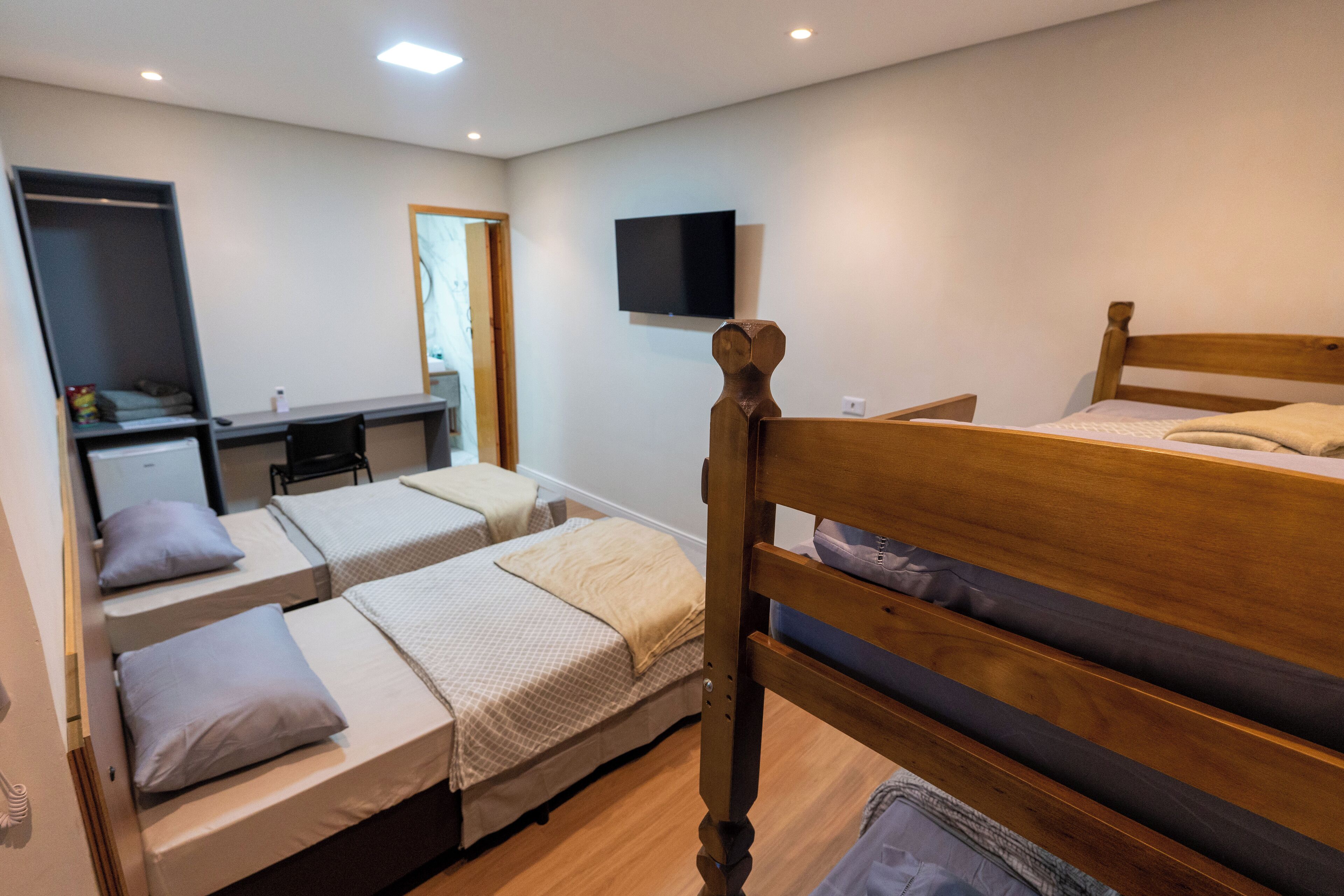 Quarto com duas camas de solteiro, uma escrivaninha, uma cadeira, televisão fixada na parede e um guarda-roupa.