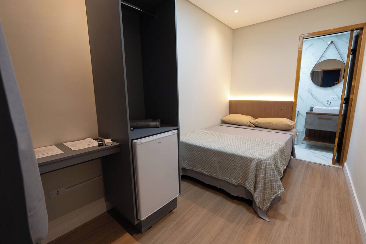 Quarto de hotel compacto com cama, pequena mesa com refrigerador, espelho e banheiro com pia e espelho.