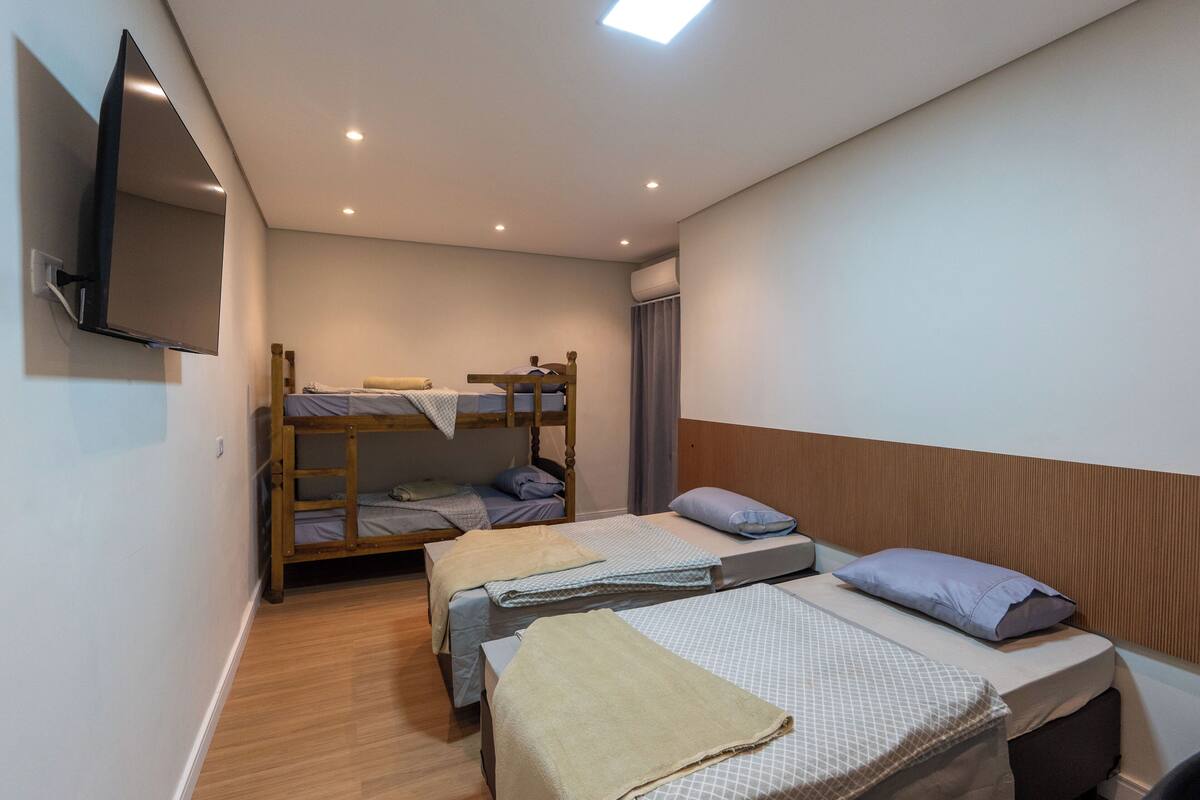 Quarto com duas camas de solteiro, uma cama plana, televisão na parede e uma luminária no teto.