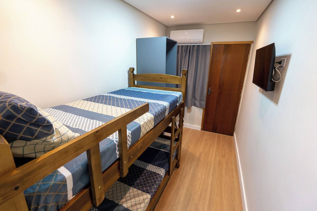 Quarto pequeno e compacto com beliche, guarda-roupa de madeira, televisão de tela plana na parede e porta de acesso a outro espaço.