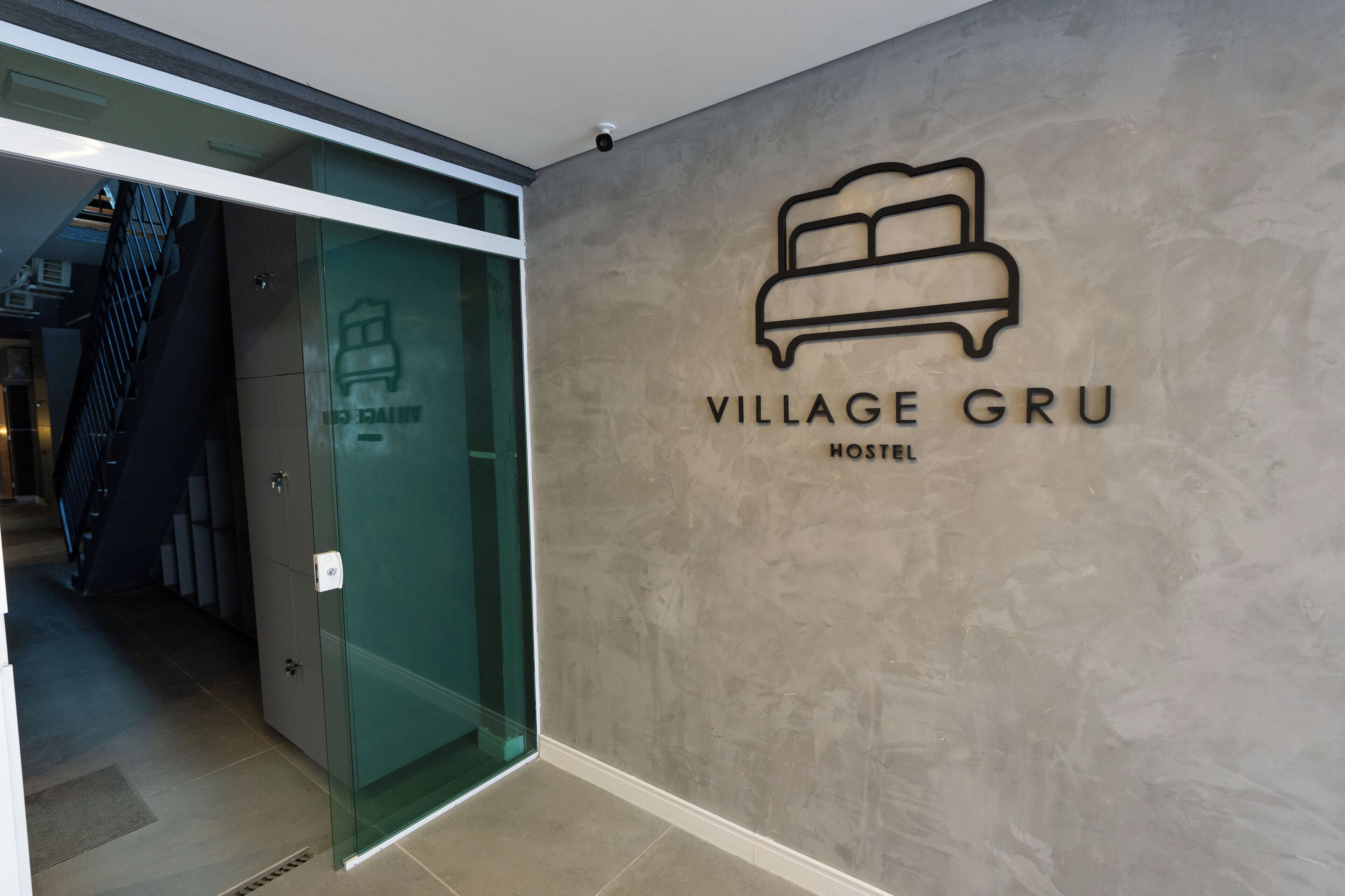 Entrada de un hostal moderno con diseño minimalista, una puerta de cristal y el logotipo de 'Village Gru Hostel' en la pared.