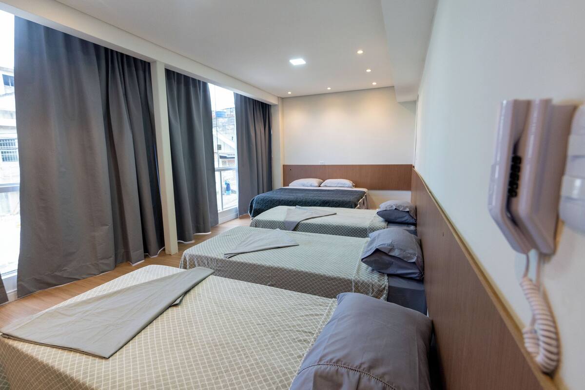 Quarto de hotel com duas camas, uma cabeceira de madeira, um telefone e uma janela com cortinas.