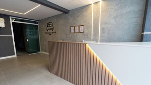 Un'area reception con scrivania in legno, una parete grigia con il cartello 'Village Gru' e una porta in vetro che dà su un'altra stanza.