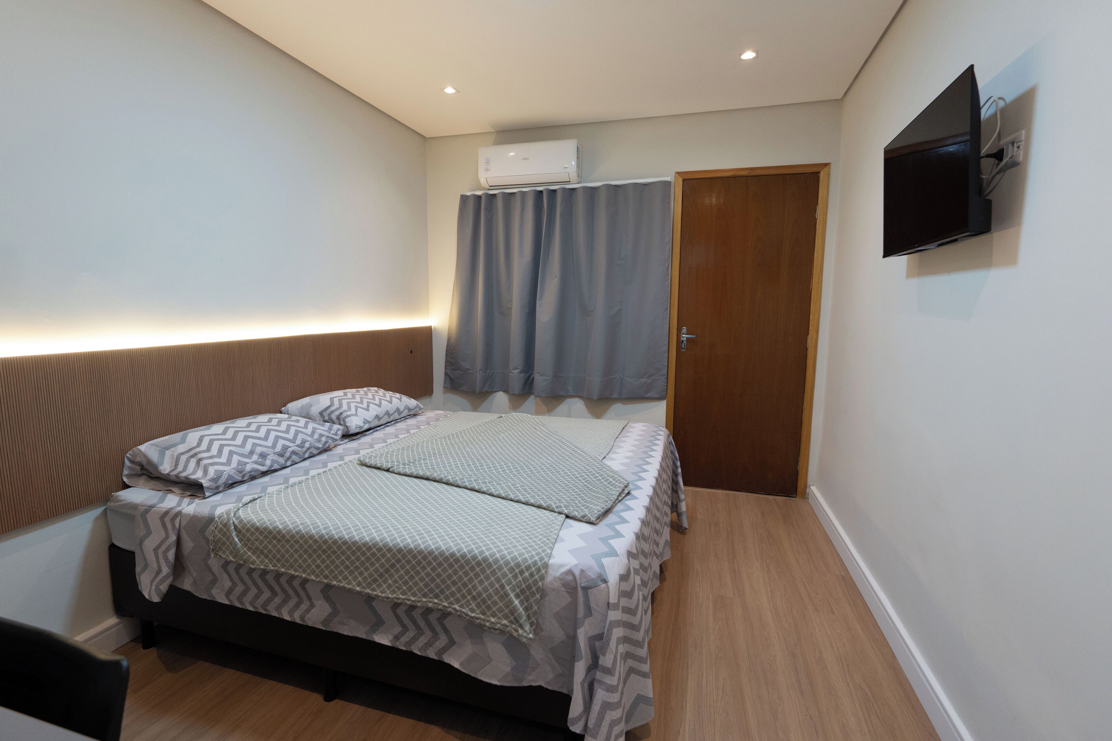A bedroom with a bed, a television mounted on the wall, and an air conditioning unit above the window.