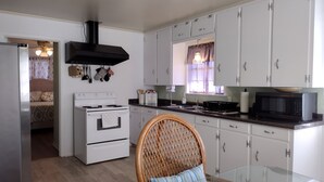 Una cocina compacta con armarios blancos, estufa, microondas y un comedor con una silla de mimbre y una mesa de vidrio.