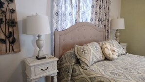 Un dormitorio con una cama, dos mesitas de noche, una mesita auxiliar, un adorno de pared y cortinas estampadas.