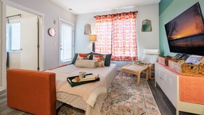 A bedroom with a bed, a chair, a TV, and a desk with books and a tray. - Le Nid Trailside 1BD Close to Beach/Downtown (Traverse City)