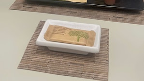 Una mesa con una botella de fragancia AD, una bandeja con servilleta de estampado de hojas verdes y dos tubos de champú de ducha.