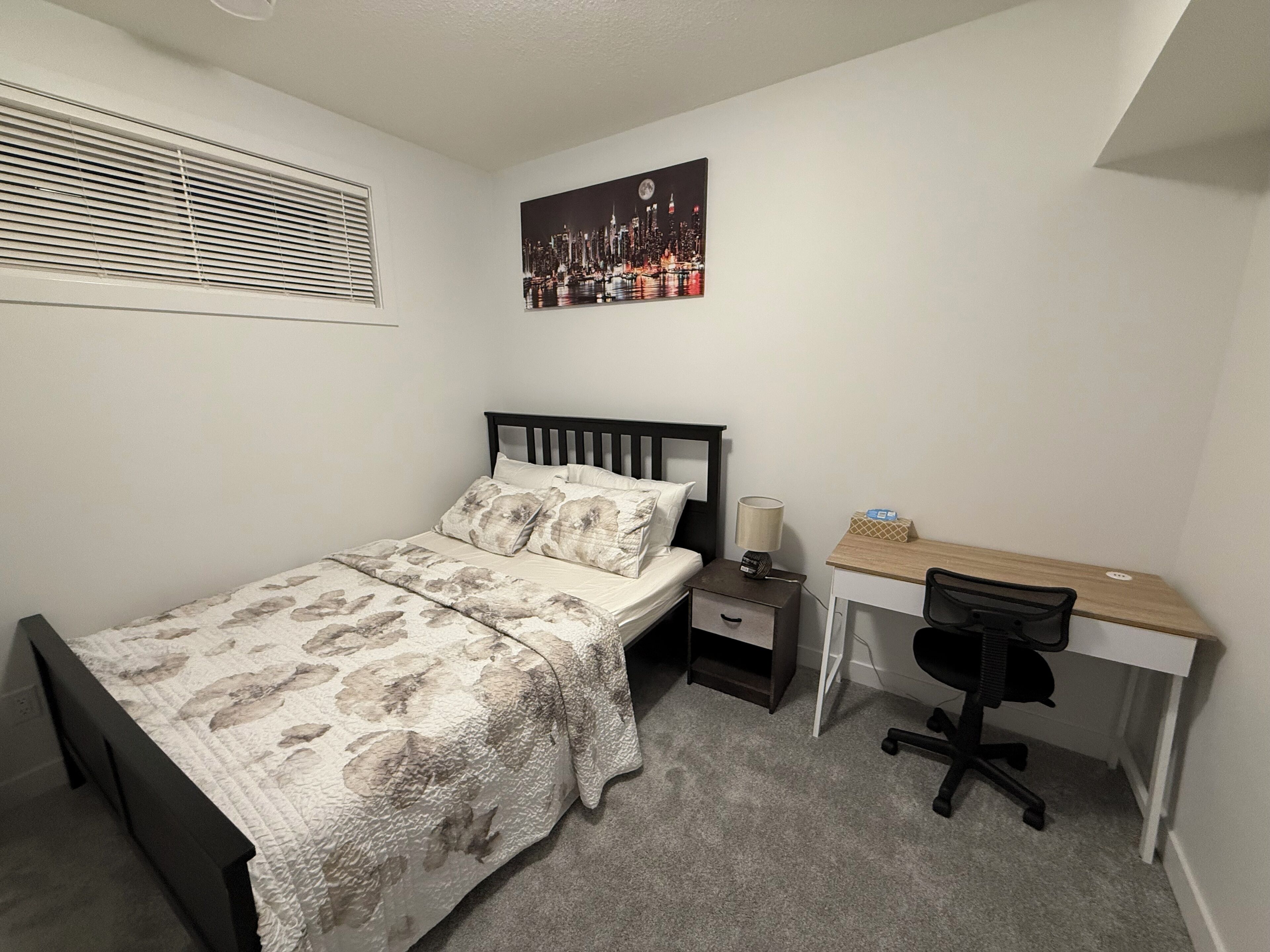 A bedroom with a bed, a desk, a chair, a nightstand, a lamp, and a cityscape painting on the wall.