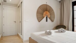 Une chambre à coucher avec un lit, deux serviettes enroulées, un ventilateur fixé au mur, une porte et une fenêtre avec des rideaux.