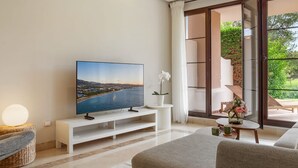 Un salon moderne doté d’un téléviseur à écran plat, d’un meuble de rangement blanc, d’un canapé et offrant une vue sur un balcon aménagé avec du mobilier d’extérieur.