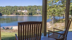 Une terrasse en bois avec une chaise berçante et une petite table, donnant sur un lac avec des maisons en arrière-plan.