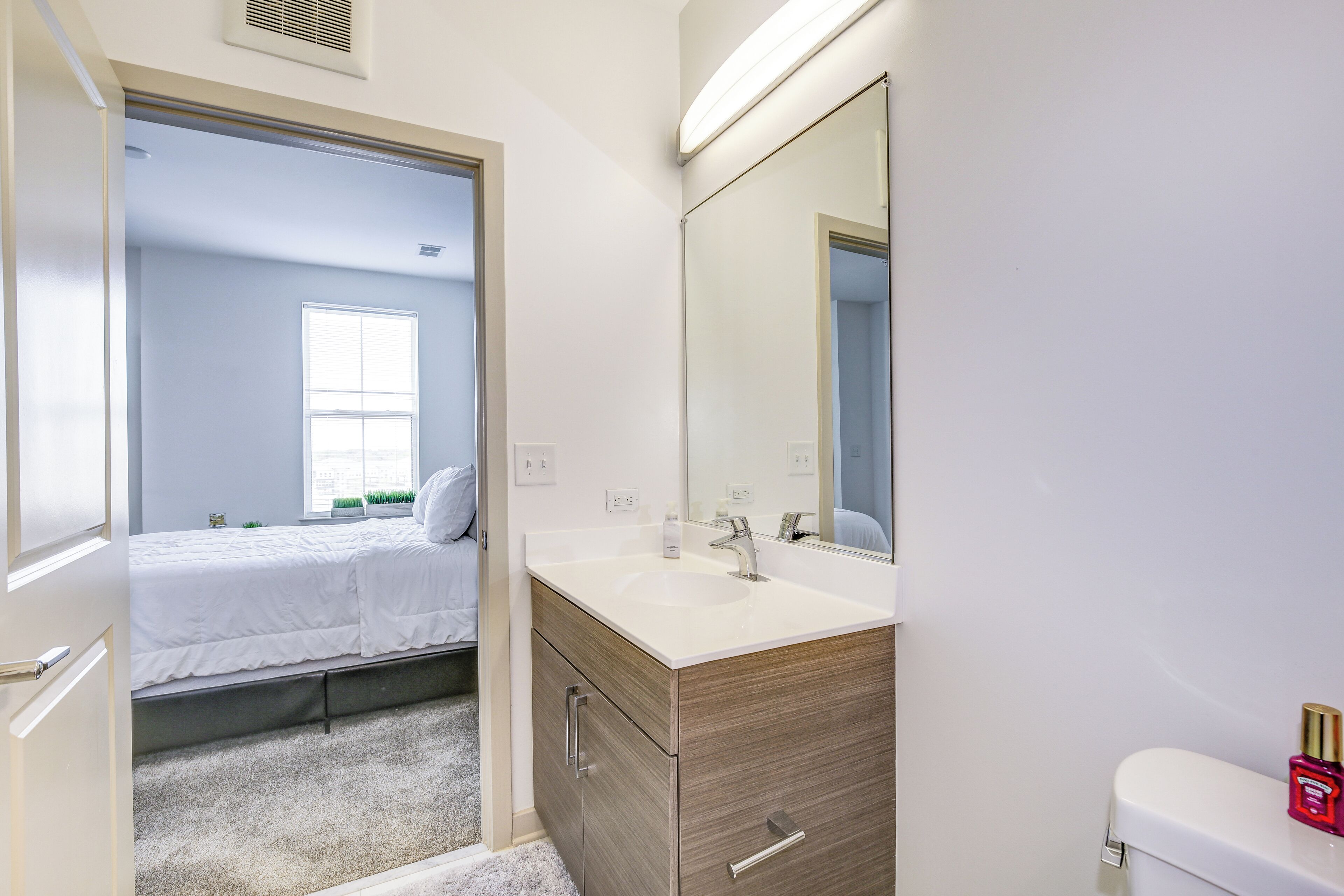 Ein Badezimmer mit einem weißen Waschbecken, einem Spiegel und einer Toilette. Ein Fenster mit Jalousien und ein Bett im angrenzenden Raum.