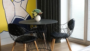 Un salon moderne avec une table ronde, deux chaises à structure métallique noire, un canapé gris et une grande fresque murale de style pop art.