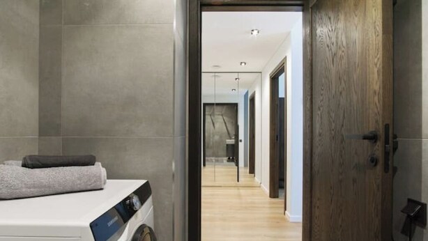 Une buanderie moderne équipée d’un lave-linge à chargement frontal, d’une porte en bois et d’un couloir menant à d’autres pièces.