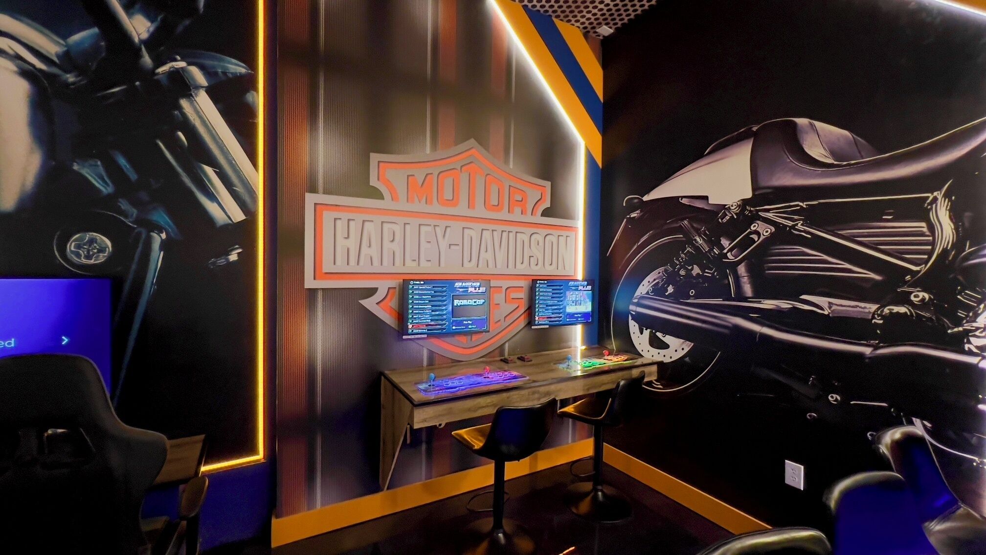 Una habitación con un adhesivo mural de una motocicleta Harley-Davidson, una configuración de juegos con dos monitores y una silla con temática de motocicleta.