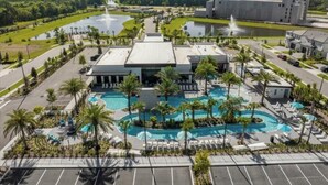 Un hotel con una amplia piscina central, palmeras y un estacionamiento.