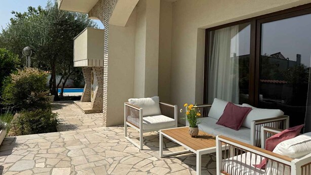 A patio with a white sofa, a wooden table, and a baby crib. There is a large window with a view of a pool and trees. - Holiday apartment with pool (Krk)