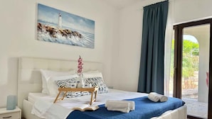 A bedroom with a large bed, a painting of a lighthouse, a view through a glass door, and a nightstand with a lamp. - Holiday apartment with pool (Krk)