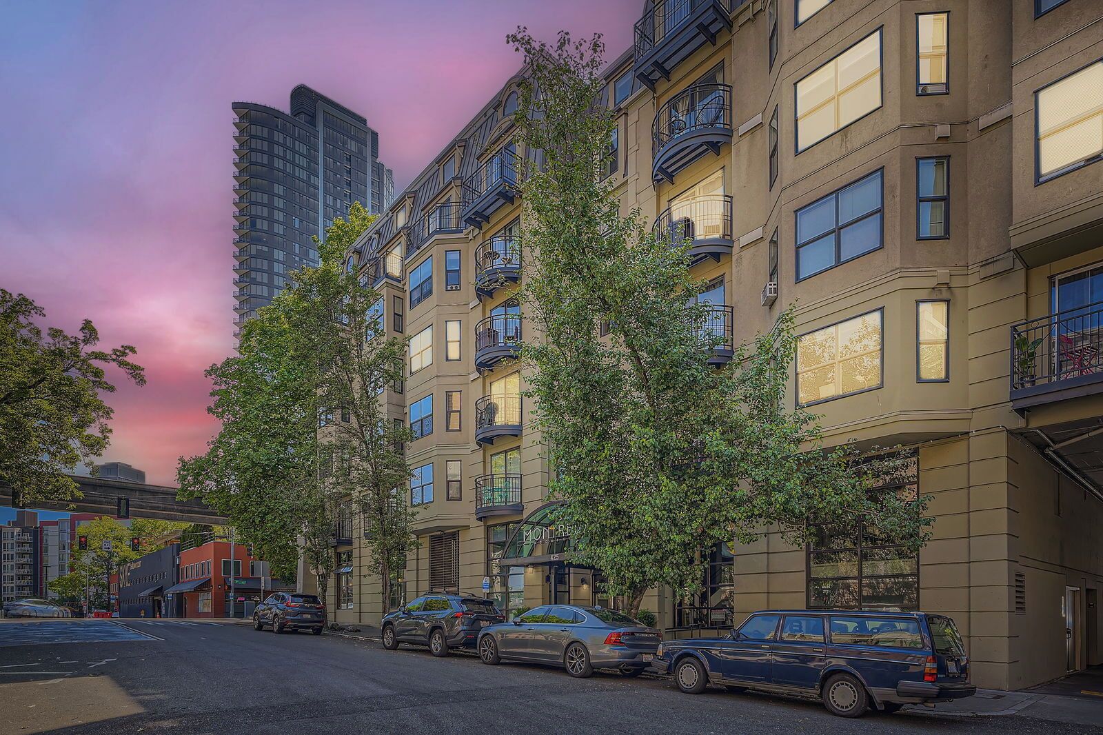 A multi-story residential building with balconies, parked cars, and trees.