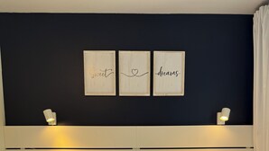 Een net opgemaakt bed met kussens en een opgevouwen handdoek, een nachtkastje en wanddecoratie met tekst.